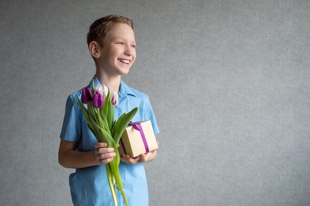 Ein süßer Junge hält einen bunten Blumenstrauß mit Tulpen und eine Schachtel mit einem Geschenk in seinen Händen