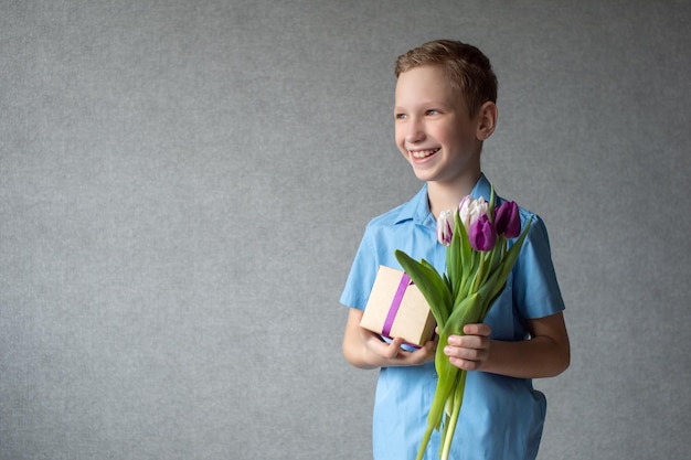 Ein süßer Junge lacht und hält einen bunten Strauß Tulpen und eine Schachtel mit einem Geschenk in den Händen