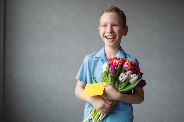 Ein süßer Junge lächelt und steht mit einem Geschenk und Tulpen und gibt einen Blumenstrauß