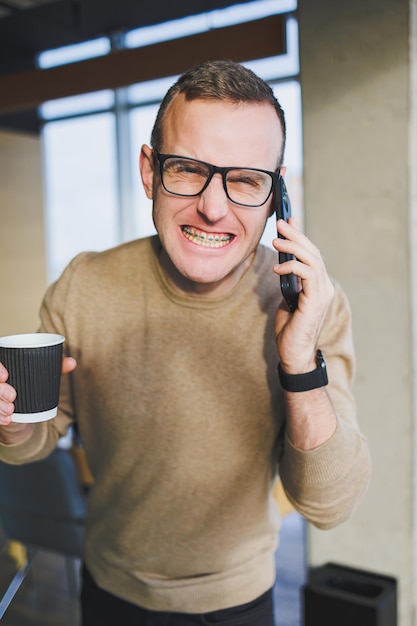 Ein süßer junger Mann in einem braunen Pullover und einer Brille spricht emotional auf einem Handy und trinkt Kaffee, während er sich im Büro entspannt Ein junger Freiberufler arbeitet aus der Ferne