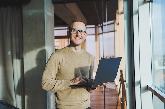 Ein süßer junger Mann in einem braunen Pullover und einer Brille steht mit einem Laptop in der Hand neben einem großen Fenster im Büro, während er im Büro arbeitet Ein junger Freiberufler arbeitet aus der Ferne