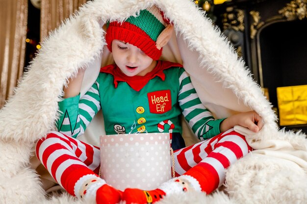 Ein süßer kleiner Junge, der als Elf verkleidet ist, öffnet ein Weihnachtsgeschenk, während er sich hinter einer flauschigen Decke in einem geschmückten Weihnachtshaus versteckt.