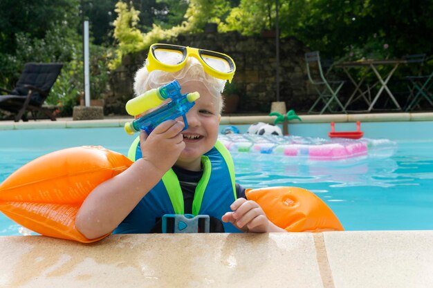 Ein süßer kleiner Junge, der Armbänder und Schutzbrillen trägt und mit einer Wasserpistole in einem Schwimmbad spielt