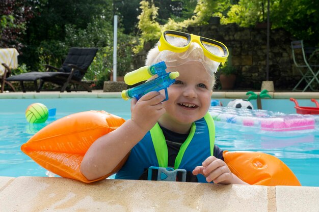 Ein süßer kleiner Junge, der Armbänder und Schutzbrillen trägt und mit einer Wasserpistole in einem Schwimmbad spielt