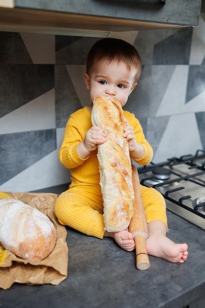 Ein süßer kleiner Junge, ein Jahr alt, sitzt und isst frisch gebackenes Roggenbrot Das Kind hält ein frisches Baguette in den Händen