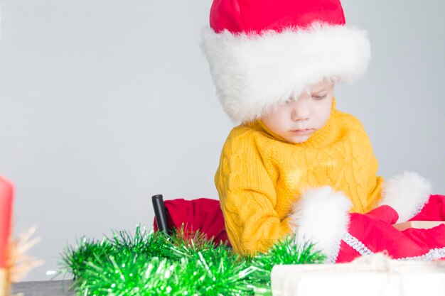 Ein süßer kleiner Junge in einem gelben Pullover, einem roten Weihnachtsmann-Hut und Weihnachtshandschuhen mit Geschenken in seinen Händen