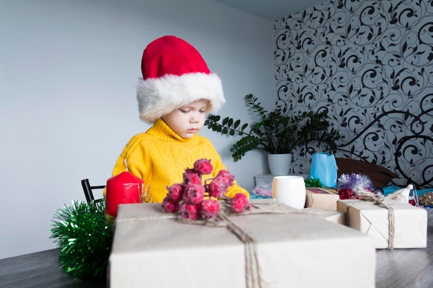 Ein süßer kleiner Junge in einem gelben Pullover und einer roten Weihnachtsmannmütze an einem Holztisch mit Geschenken, die in Kraftpapier verpackt sind