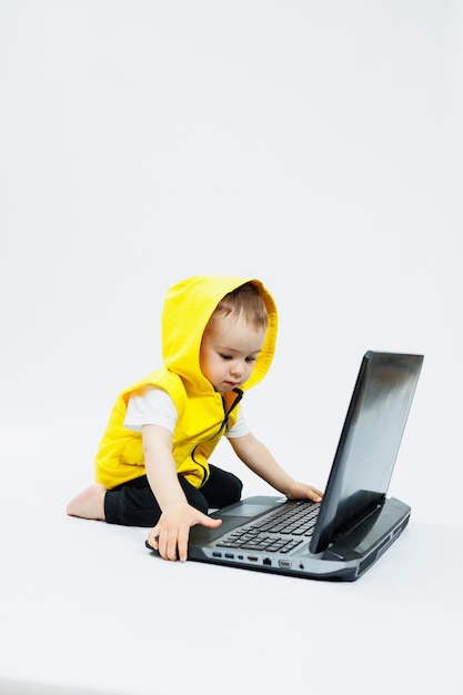 Ein süßer kleiner Junge in einer gelben Weste sitzt an einem digitalen Laptop auf weißem Hintergrund. Kind und modernes tragbares Computergerät