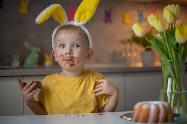 Ein süßer kleiner Junge mit Hasenohren am Ostertag isst einen Schokoladen-Osterhasen Ein Kind spielt Eiersuche für Ostern Charmantes Kind feiert Ostern zu Hause