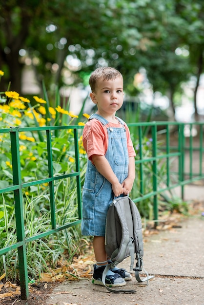 Ein süßer kleiner Junge schaut in die Kamera und hält eine Tasche in den Händen, gekleidet in Jeans-Overalls