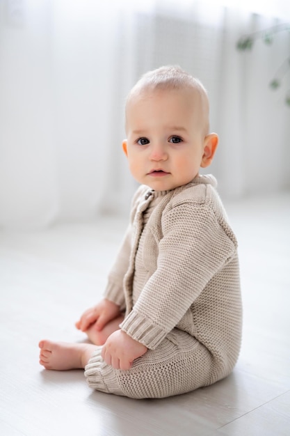 Ein süßer kleiner Junge sitzt auf dem Boden und lernt, ein großes Porträt eines Kindes mit braunen Augen und blonden Haaren in einem Strickanzug in Pastellfarben zu sitzen