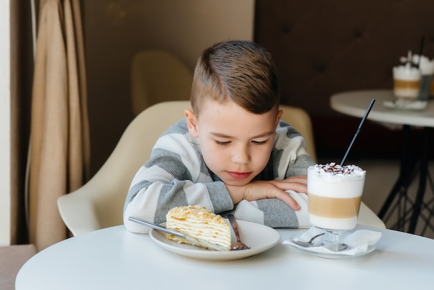 Ein süßer kleiner Junge sitzt in einem Café und betrachtet eine Nahaufnahme von Kuchen und Kakao.