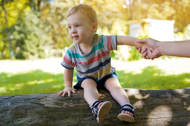 Foto ein süßer kleiner junge von anderthalb jahren sitzt auf einem baumstamm und hält die hand seines vaters im park an einem sonnigen sommertag glücklich lächelndes kleinkind selektiver fokus