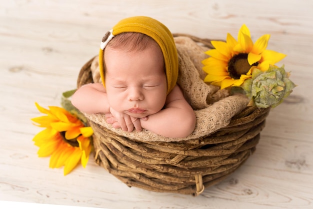 Ein süßer neugeborener Junge schläft in den ersten Lebenstagen mit einem gelb-orangefarbenen Hut vor einem Hintergrund aus hellweißem Holz in einem geflochtenen Holzkorb mit Sonnenblumenblüten. Professionelles Fotografie-Porträt