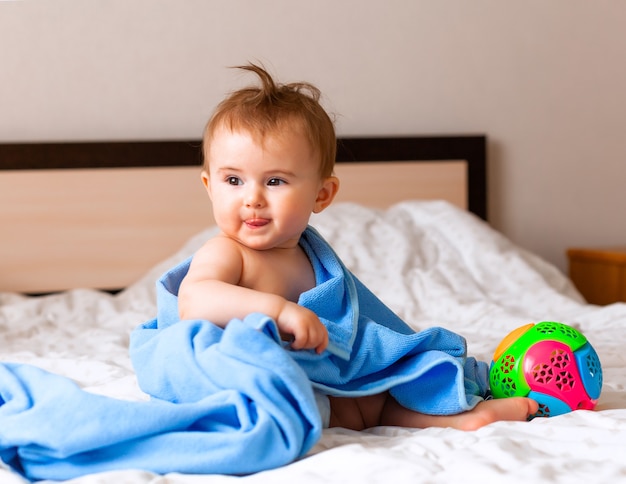 Ein süßes blondes Baby, das in ein blaues Badetuch gewickelt ist, spielt mit einem Ball, während es auf dem Bett im Schlafzimmer sitzt. glückliches 6 Monate altes Baby, das mit Ball spielt