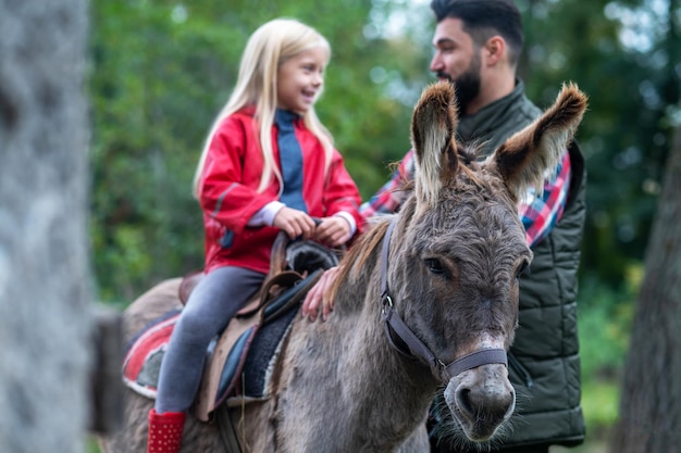 Ein süßes blondes Mädchen, das einen Esel auf einer Farm reitet, ihr Vater hilft ihr