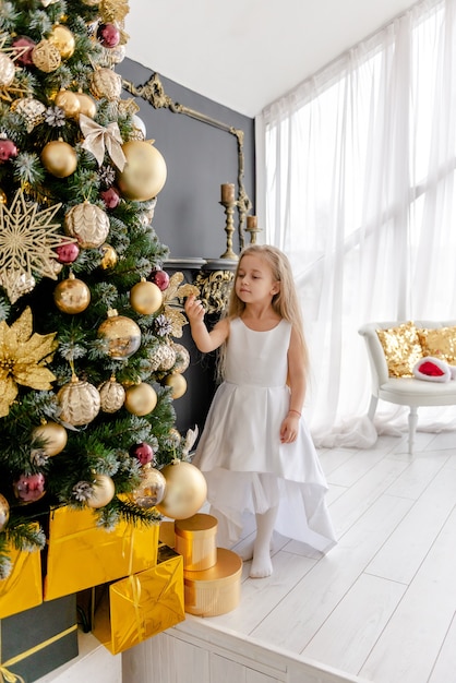 Ein süßes blondes Mädchen schmückt vor Weihnachten zu Hause einen Baum. Glückliche Kindheit. Einen Weihnachtsbaum vor Weihnachten schmücken.