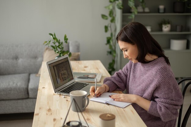 Ein süßes brünettes Mädchen in einem lila Pullover sitzt mit einem Laptop an einem Tisch und macht sich Notizen