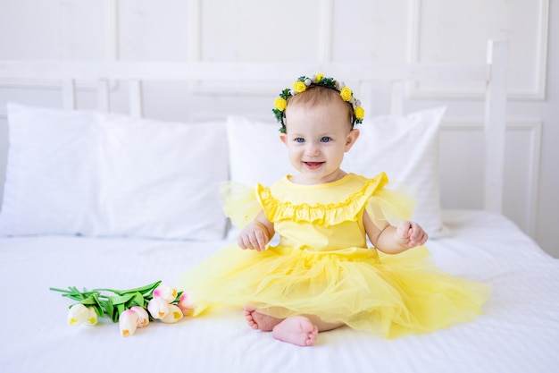 Ein süßes, gesundes Mädchen in einem gelben Kleid mit rosafarbenen Tulpenblumen sitzt auf einem Bett auf weißer Bettwäsche und blickt in die Kamera