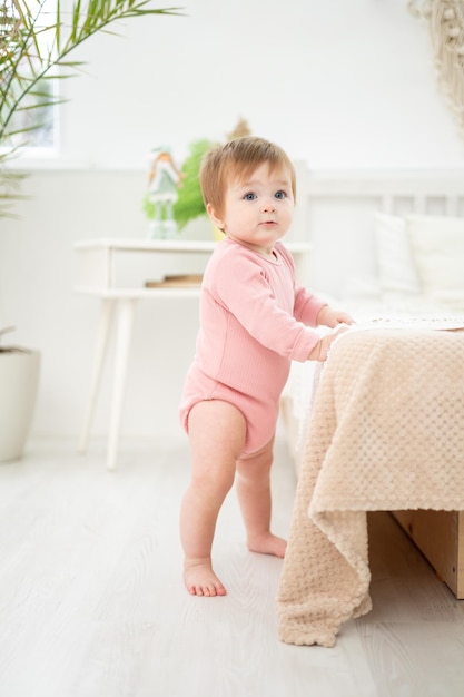 Ein süßes, gesundes Mädchen steht am Bett im Schlafzimmer zu Hause, die ersten Schritte des Babys, das Baby lernt zu laufen