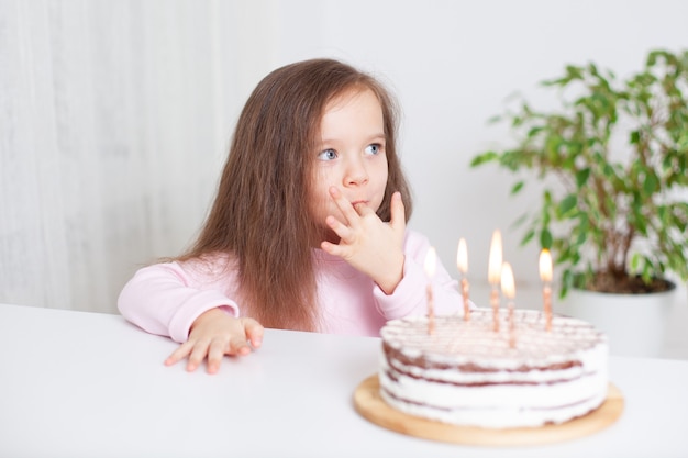 Ein süßes glückliches kleines Mädchen mit blonden Haaren und blauen Augen leckt die Sahne mit einem Fingergeburtstag