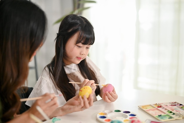 Ein süßes junges asiatisches Mädchen konzentriert sich auf das Malen von Ostereiern mit Wasserfarben Glückliche Familie und Urlaub