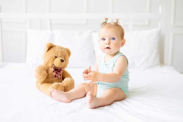 Ein süßes kleines Baby in einem Body aus natürlichem Baumwollstoff sitzt auf weißer Bettwäsche im Schlafzimmer Ein glückliches kleines Baby im Bett Textilien und Bettwäsche für Kinder