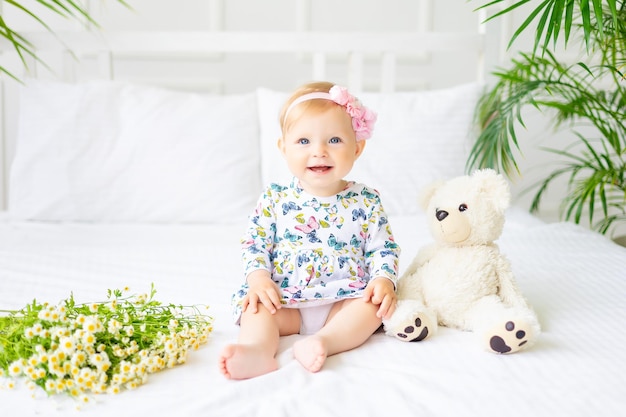 Ein süßes kleines Baby in einem Kleid aus natürlichem Baumwollstoff sitzt auf weißer Bettwäsche im Schlafzimmer Ein kleines glückliches Kind im Bett mit einem Teddybären und einem Strauß Gänseblümchen