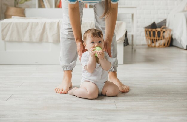 Ein süßes kleines Baby von sechs Monaten sitzt auf dem Boden neben Mamas Beinen und lächelt.