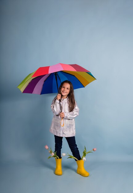 Ein süßes kleines brünettes Mädchen in Gummistiefeln mit Blumen steht unter einem bunten Regenschirm auf blauem Hintergrund mit Platz für Text