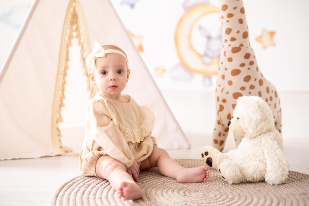 Ein süßes kleines europäisches Mädchen mit braunen Augen in einem beigen Body aus natürlichem Stoff spielt im heimischen Kinderzimmer vor dem Hintergrund eines Wigwams und Plüschspielzeug