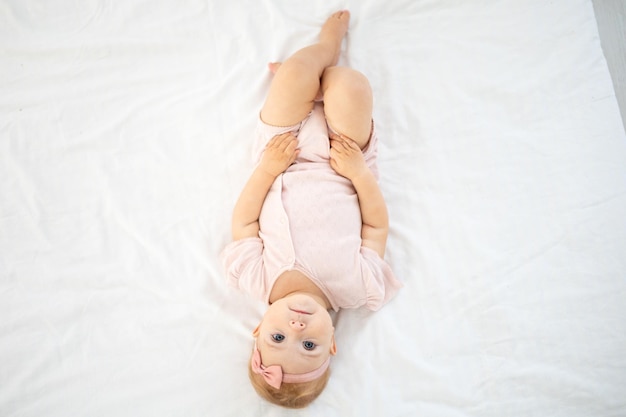 Ein süßes kleines gesundes Mädchen bis zu einem Jahr in einem rosa Body aus natürlichem Stoff liegt auf dem Bett auf weißer Bettwäsche im Schlafzimmer und schaut auf die Kamera, das Baby ist zu Hause