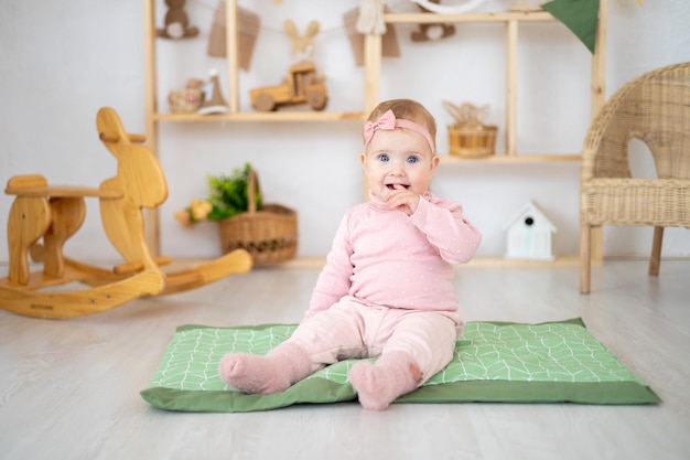 Ein süßes kleines gesundes Mädchen bis zu einem Jahr in einem rosafarbenen Anzug aus natürlichem Stoff sitzt auf einem Teppich in einem Kinderzimmer mit hölzernen Lernspielzeugen, die lächelnd auf die Kamera schauen