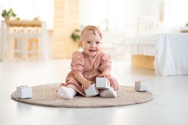 Ein süßes kleines gesundes Mädchen bis zu einem Jahr in einem rosafarbenen Kleid aus natürlichem Stoff sitzt auf einem Teppich im Wohnzimmer des Hauses und spielt mit Holzwürfeln die Entwicklung des Kindes zu Hause