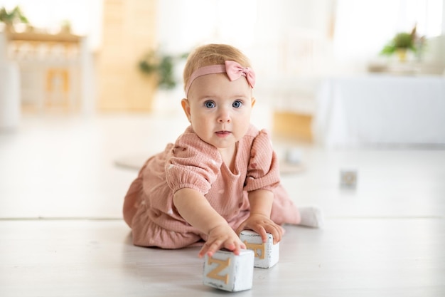 Ein süßes kleines gesundes Mädchen bis zu einem Jahr in einem rosafarbenen Kleid aus natürlichem Stoff sitzt auf einem Teppich im Wohnzimmer des Hauses und spielt mit Holzwürfeln die Entwicklung des Kindes zu Hause