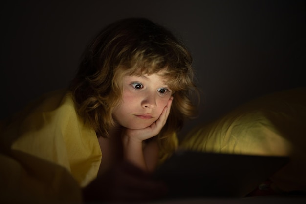 Ein süßes kleines Kind liegt unter einer Decke mit einem Tablet und überrascht ein Kind im Bett unter einer Dekke, das ein Video sieht.