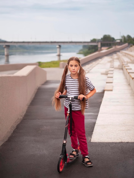Ein süßes kleines Mädchen fährt mit dem Roller auf der Brücke Außenporträt