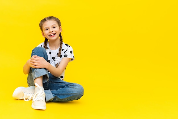 Ein süßes kleines Mädchen in Blue Jeans sitzt auf einem gelb isolierten Hintergrund und lächelt