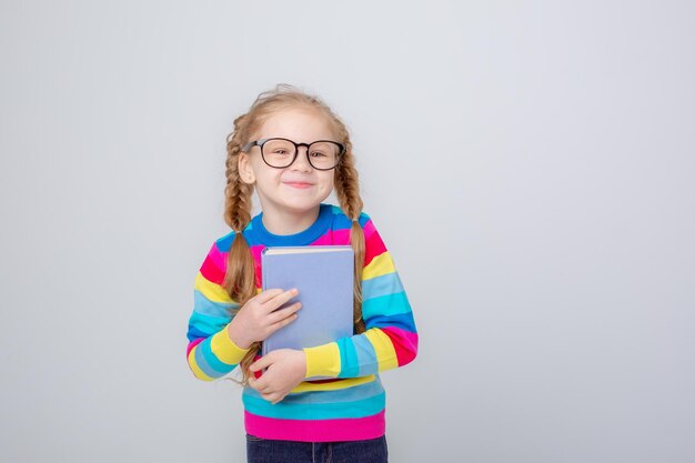 Ein süßes kleines Mädchen in einem bunten Pullover und einer Brille hält ein Buch auf weißem Hintergrund und lächelt