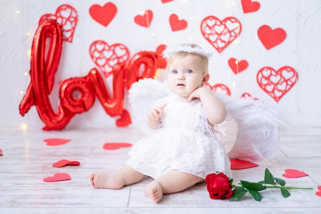 Ein süßes kleines Mädchen in einem Engelskostüm mit Flügeln auf einem Hintergrund aus roten Herzen und der Inschrift lieben das Konzept des Valentinstags