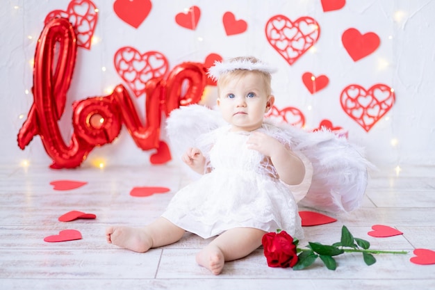 Ein süßes kleines Mädchen in einem Engelskostüm mit Flügeln auf einem Hintergrund aus roten Herzen und der Inschrift lieben das Konzept des Valentinstags