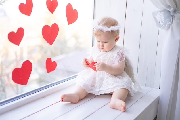 Ein süßes kleines Mädchen in einem Engelskostüm mit Flügeln sitzt auf der Fensterbank neben dem Fenster vor dem Hintergrund roter Herzen Valentinstag Konzept Valentinstag