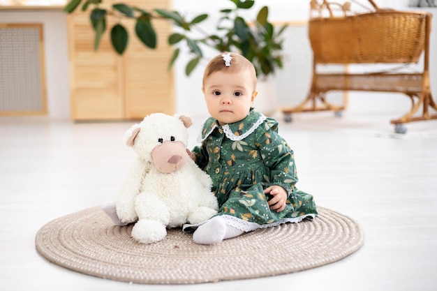 Ein süßes kleines Mädchen in einem grünen Baumwollkleid sitzt auf einem Teppich im Wohnzimmer des Hauses und schaut lächelnd in die Kamera und spielt mit einem weißen Teddybären