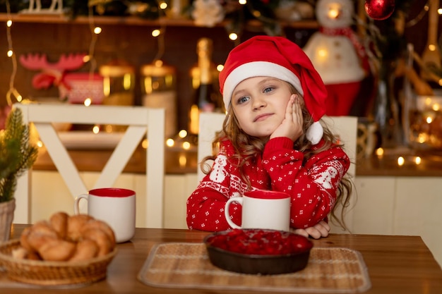 Ein süßes kleines Mädchen in einem roten Pullover und einer Weihnachtsmannmütze ist in der Küche, trinkt Tee mit einer Torte und wartet auf das neue Jahr oder Weihnachten