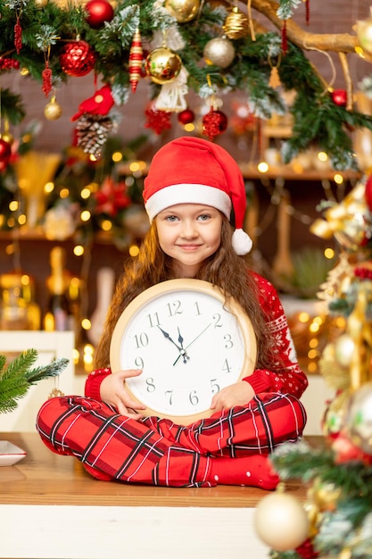 Ein süßes kleines Mädchen in einer Weihnachtsmannmütze und einem roten Pullover mit einer Uhr in den Händen lacht vor Glück in der dunklen Küche des Hauses in Erwartung des neuen Jahres und Weihnachten