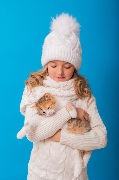 Ein süßes kleines Mädchen in einer weißen Strickmütze und einem Pullover hält ein Kätzchen auf blauem Hintergrund im Studio Platz für Text Das Konzept von Weihnachten ist das Symbol des Jahres