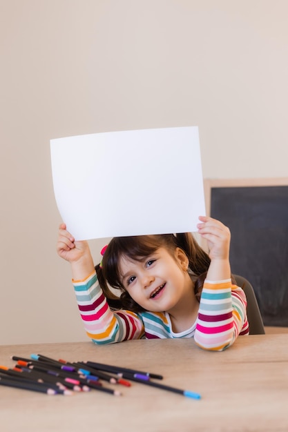 Ein süßes kleines Mädchen in einer Zeichenstunde hält ein leeres weißes Blatt in ihren Händen, Platz für Text Kinder und Kreativität