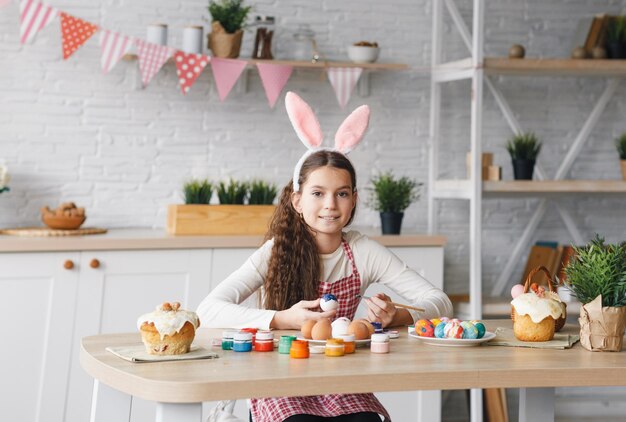 Ein süßes kleines Mädchen in Hasenohren bereitet bemalte Ostereier für Ostern vor, die am Küchentisch sitzen. Frohe Ostern