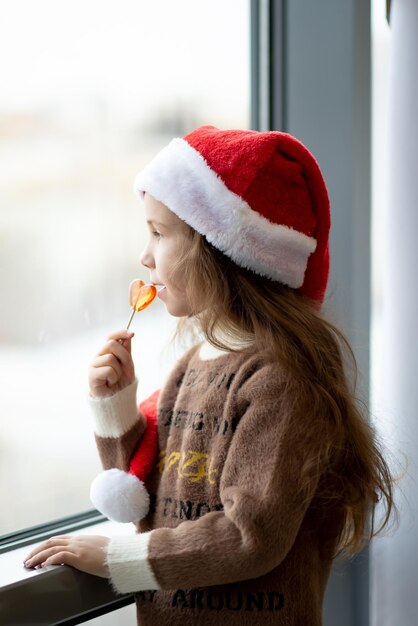 Ein süßes kleines Mädchen in Santa's Hut hält eine Süßigkeit in Form eines Herzens in ihren Händen