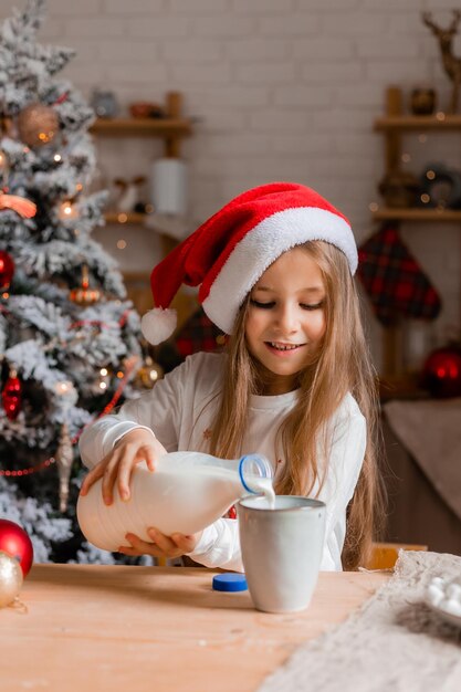 ein süßes kleines Mädchen in Weihnachtsmannshut und Pyjama trinkt Milch in der Küche für Weihnachten Mockup